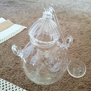 Könnte beinhalten: Eine klare Glas-Bowle mit Deckel und Schöpflöffel. Die Schüssel hat eine runde Form mit einem strukturierten Muster. Der Deckel ist gewölbt mit vertikalen Rippen. Der Schöpflöffel hat einen langen Griff und eine runde Kelle.