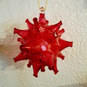May include: A vibrant red glass ornament with a spiky, starburst design. The ornament has a clear glass loop for hanging and is suspended by a gold string. The spiky design gives the ornament a unique, eye-catching appearance.