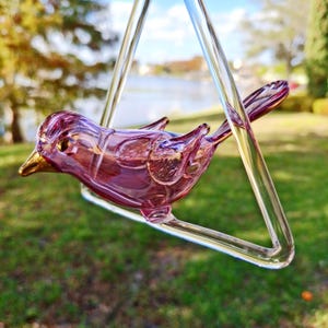 Puede incluir: Una escultura de pájaro de cristal hecha a mano en tonos morados y dorados, posada en un columpio de cristal transparente. El pájaro tiene alas detalladas y un pico dorado. El columpio está suspendido contra un fondo borroso al aire libre.