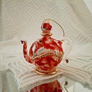 Puede incluir: Adorno decorativo de tetera de cristal con diseño veteado rojo y blanco. La tetera tiene base, ribete y asa dorados. Se adjunta una cuerda dorada para colgarla. La tetera mide aproximadamente 10 cm de altura.