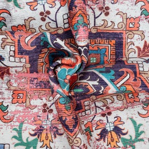Puede incluir: Primer plano de una tela doblada con un patr&oacute;n ornamentado y desgastado. El dise&ntilde;o presenta una mezcla de colores, incluyendo naranja, turquesa, morado y rosa, sobre un fondo blanco. El patr&oacute;n se asemeja a una alfombra o tapiz tradicional.