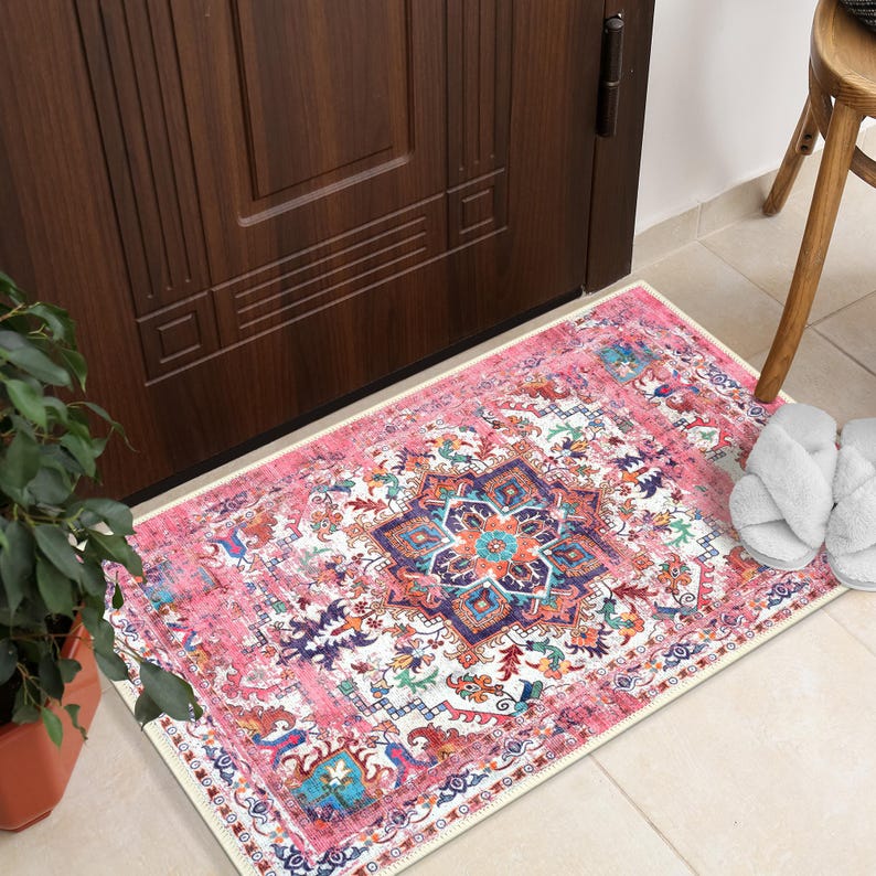 Puede incluir: Una alfombra rectangular con estampado rosa y blanco con un dise&ntilde;o de medall&oacute;n central. La alfombra presenta un aspecto envejecido y vintage con un borde e intrincados patrones florales y geom&eacute;tricos. Unas zapatillas blancas descansan sobre el suelo embaldosado.