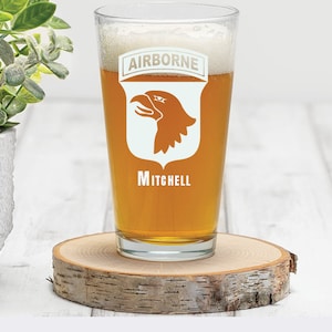 May include: A clear pint glass with the word "AIRBORNE" above a shield with an eagle head and the name "Mitchell" below. The glass is filled with a light-colored liquid and rests on a wooden coaster.