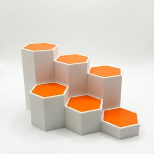 May include: A tiered display stand featuring six white hexagonal pillars of varying heights, each topped with a vibrant orange hexagonal surface. The structure is arranged in a stepped formation, creating a multi-level platform for showcasing items.