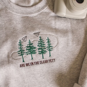 May include: A grey sweatshirt with an embroidered design of three pine trees and paper aeroplanes with the text "Are we in the clear yet?"