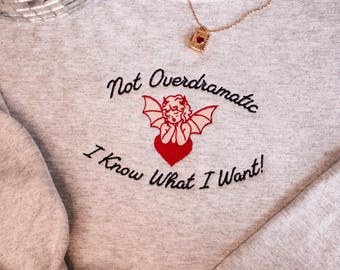 Puede incluir: Sudadera gris con el texto "Not Overdramatic I Know What I Want!" bordado en la parte delantera. El texto está en negro y hay un corazón rojo y un pequeño ángel con alas bordado encima del texto.