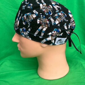 May include: A black surgical scrub cap featuring a repeating pattern of astronauts in white, blue, and orange spacesuits. The cap has a black tie closure and a small white tag with text.