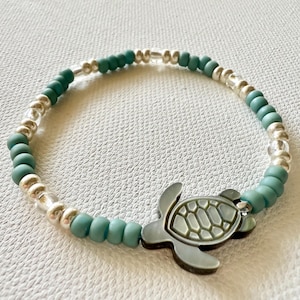 Op de afbeelding: Een armband met een zeeschildpadbedel. De armband heeft afwisselend turquoise en zilveren kralen. De zeeschildpadbedel heeft een gedempte grijsgroene kleur met een gestructureerd schildontwerp. De armband ligt op een witte ondergrond.