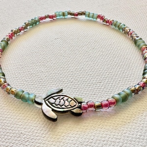 May include: Anklet featuring a silver-tone turtle charm and a beaded strand. The beads are in shades of green, pink, and iridescent gold. The anklet is designed to be worn around the ankle.