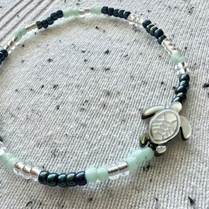 May include: A beaded bracelet featuring a central sea turtle charm made of iridescent shell material. The band consists of alternating small round beads in shades of pale mint green, metallic dark charcoal, and clear glass, arranged in a repeating pattern.