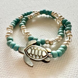 May include: Two beaded bracelets. One bracelet features silver beads, the other has turquoise beads. A silver turtle charm is attached to the turquoise bracelet. The bracelets are on a white surface.