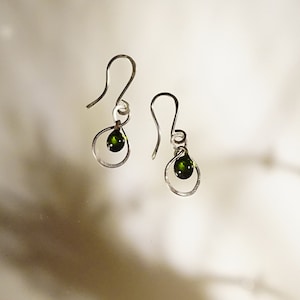 May include: A pair of silver earrings with green gemstone accents. The earrings feature a simple, delicate design with a small, round gemstone hanging from a curved wire.