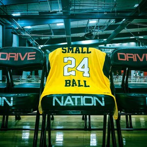 Puede incluir: Una camiseta de baloncesto amarilla con ribetes negros y las palabras "SMALL BALL" y el n&uacute;mero "24" en la espalda. La camiseta est&aacute; colgada en el respaldo de una silla negra con la palabra "DRIVE" en la espalda.