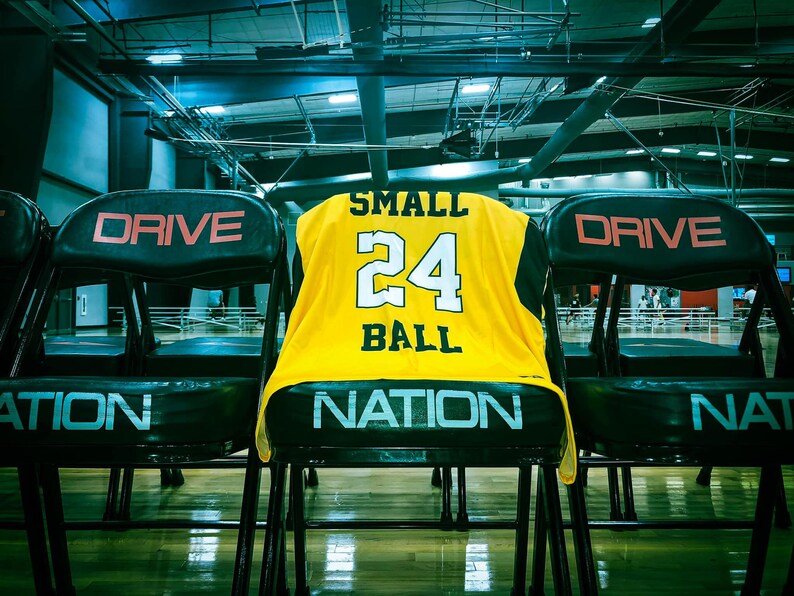 Puede incluir: Una camiseta de baloncesto amarilla con ribetes negros y las palabras "SMALL BALL" y el n&uacute;mero "24" en la espalda. La camiseta est&aacute; colgada en el respaldo de una silla negra con la palabra "DRIVE" en la espalda.