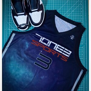 Puede incluir: Una camiseta de baloncesto negra con estampado de galaxia y el texto "Toney Sports 3" en blanco y rojo. La camiseta tiene un ribete blanco alrededor del cuello y las sisas.