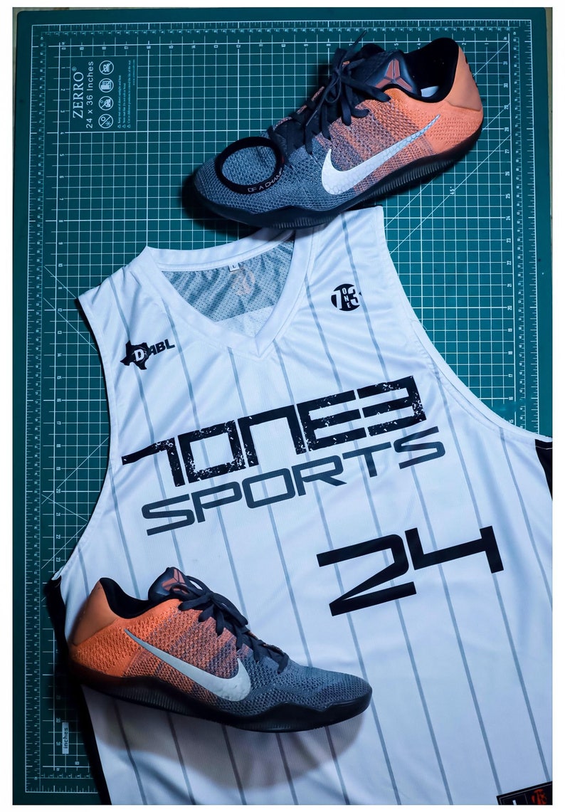 Puede incluir: Camiseta de baloncesto blanca con rayas negras y el texto "ONE3 SPORTS 24" en la parte delantera. Dos zapatillas de baloncesto negras, naranjas y grises est&aacute;n a cada lado de la camiseta.