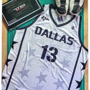 Puede incluir: Camiseta de baloncesto blanca con ribetes negros y un patr&oacute;n de estrellas. La camiseta tiene el texto "DABL" y "Dallas" en la parte delantera, junto con el n&uacute;mero "13".