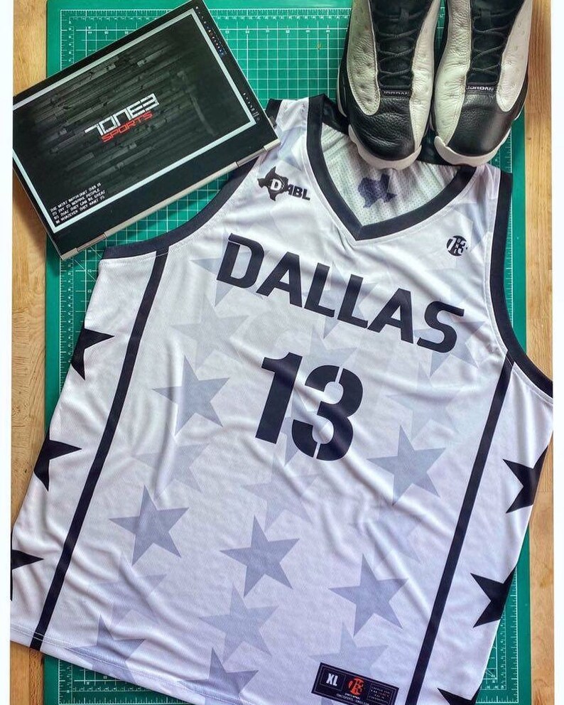 Puede incluir: Camiseta de baloncesto blanca con ribetes negros y un patr&oacute;n de estrellas. La camiseta tiene el texto "DABL" y "Dallas" en la parte delantera, junto con el n&uacute;mero "13".