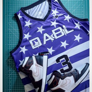 Puede incluir: Una camiseta de baloncesto de color morado y blanco con estrellas y rayas. La camiseta tiene el texto "DABL" en la parte delantera y el n&uacute;mero "3" en la parte trasera. Dos zapatillas negras y blancas est&aacute;n encima de la camiseta.