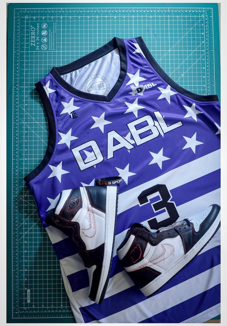 Puede incluir: Una camiseta de baloncesto de color morado y blanco con estrellas y rayas. La camiseta tiene el texto "DABL" en la parte delantera y el n&uacute;mero "3" en la parte trasera. Dos zapatillas negras y blancas est&aacute;n encima de la camiseta.