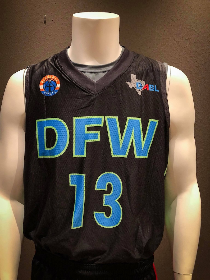Puede incluir: Camiseta de baloncesto negra con un "DFW 13" verde y azul en la parte delantera. La camiseta tiene un peque&ntilde;o logotipo en el lado izquierdo que dice "Apogee Sports" y un logotipo m&aacute;s grande en el lado derecho que dice "D-ABL" con un contorno de Texas.
