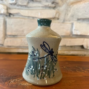 May include: A small, bell-shaped ceramic vessel with a speckled beige glaze. The body features a hand-painted design of a dark blue dragonfly hovering over green grass blades with small white dots, topped with a dark green glazed neck opening.
