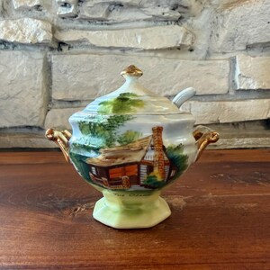 Puede incluir: Un azucarero de porcelana con tapa y dos asas doradas. El cuenco presenta una escena pintada a mano de una cabaña con chimenea, árboles verdes y el texto The Ozarks. Tiene una base de pedestal y una pequeña abertura en la tapa para cuchara.