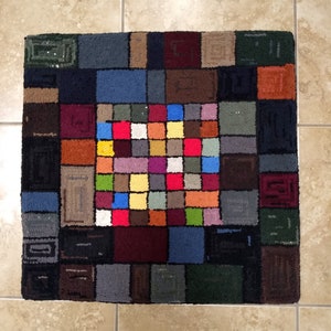 May include: A colorful rug with a geometric pattern of squares in various shades of red, blue, green, yellow, brown, and gray. The rug is made of a soft, plush material and has a unique, modern design.