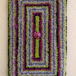 May include: A rectangular rug with a multi-colored design. The rug is made of fabric strips and has a black, green, gray, and purple color scheme. A pink flower with a rhinestone center is in the center of the rug.