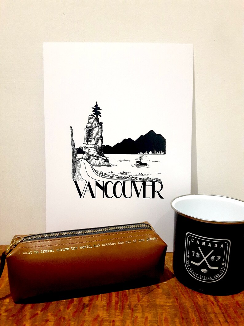 May include: A black and white illustration of a rocky island with a tree on top, a boat in the water, and the word "Vancouver" printed below. The illustration is on a white piece of paper, which is sitting on a wooden table. There is a brown leather pencil case with the text "I want to travel across the world, and breathe the air of new places" printed on it. There is also a black and white mug with the text "CANADA 1867" printed on it.