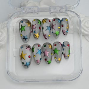 Pode incluir: Um conjunto de unhas postiças transparentes em forma oval. Cada unha apresenta um design de estrelas coloridas em tons de amarelo, verde, azul e rosa, com pequenos enfeites redondos vermelhos e azuis. As unhas são exibidas em uma caixa de plástico transparente.