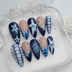 May include: A set of ten almond-shaped false nails in a clear plastic case. The nails feature a navy blue base with white star and dot designs, and some have a nude-colored base with blue accents. Each nail is adorned with a small, round, blue gem.