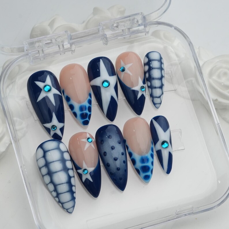 May include: A set of ten almond-shaped false nails in a clear plastic case. The nails feature a navy blue base with white star and dot designs, and some have a nude-colored base with blue accents. Each nail is adorned with a small, round, blue gem.