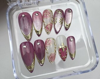 May include: A set of ten artificial nails in a clear plastic case. The nails feature a variety of designs, including a gradient of pink and gold, floral patterns, and gold accents. Some nails have a small rhinestone.