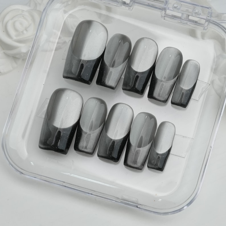 May include: A set of ten gray and white press-on nails in a clear plastic case. The nails have a glossy finish with a gradient design, transitioning from a dark gray at the base to a lighter gray and white at the tip.
