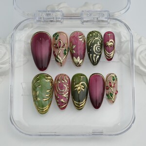 May include: A set of ten artificial nails in a clear plastic case. The nails feature a variety of designs, including burgundy, green, and pink hues, with gold floral and swirl accents. The nails are oval-shaped.