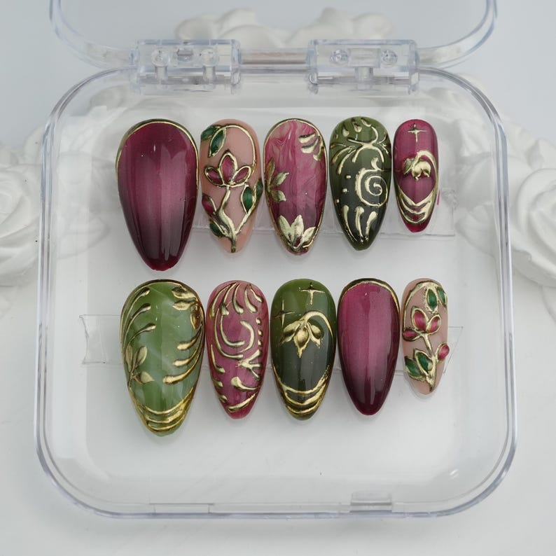 May include: A set of ten artificial nails in a clear plastic case. The nails feature a variety of designs, including burgundy, green, and pink hues, with gold floral and swirl accents. The nails are oval-shaped.