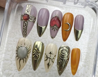 Pode incluir: Um conjunto de dez unhas postiças com diferentes designs. As unhas são todas longas e pontiagudas, e têm uma variedade de cores e padrões. Alguns dos designs incluem uma cruz, um coração, uma lua crescente, um sol e uma rosa. As unhas estão dispostas em uma caixa de plástico transparente.