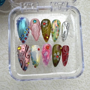 Könnte beinhalten: Eine transparente Kunststoffbox mit zehn künstlichen Nägeln. Die Nägel sind in verschiedenen Farben gehalten, darunter Blau, Rosa, Grün und Silber, und sind mit Strasssteinen, Glitzer und aufwendigen Designs verziert. Die Nägel sind mandelförmig.