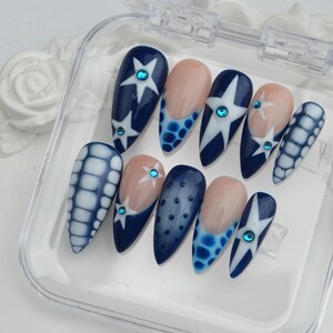 May include: A set of ten artificial nails in a clear plastic case. The nails feature various designs, including navy blue with white star patterns, light blue polka dots, and a light pink base with a blue border. Each nail has a small, round, blue gem.
