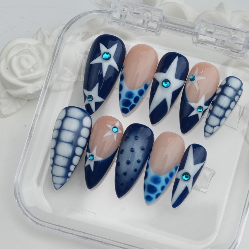 May include: A set of ten artificial nails in a clear plastic case. The nails feature various designs, including navy blue with white star patterns, light blue polka dots, and a light pink base with a blue border. Each nail has a small, round, blue gem.