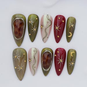 May include: A set of ten almond-shaped press-on nails featuring a celestial theme. The designs include olive green, deep red, and cream bases adorned with gold starbursts, crescent moons, small gold studs, and raised silver-toned wavy lines.
