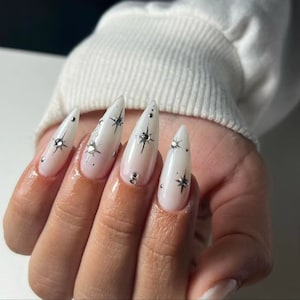 Puede incluir: Uñas blancas en forma de stiletto con efecto degradado y detalles de estrellas plateadas.