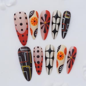 Op de afbeelding: Een set van tien amandelvormige kunstnagels met diverse ontwerpen, waaronder zwarte stippen op een rode en huidkleurige basis, oranje bloemmotieven op crème, rode sterpatronen, zwarte geometrische rasters en wit-oranje vierkante omlijningen.