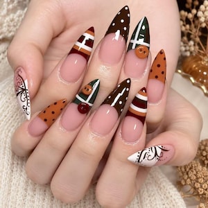 Op de afbeelding: Puntige, amandelvormige nagels in een French manicure-stijl met een nude basis. De uiteinden zijn versierd met bruine, witte en groene patronen, waaronder stippen, strepen en ruiten. Kleine, ronde, driedimensionale knoopjes zijn bevestigd.