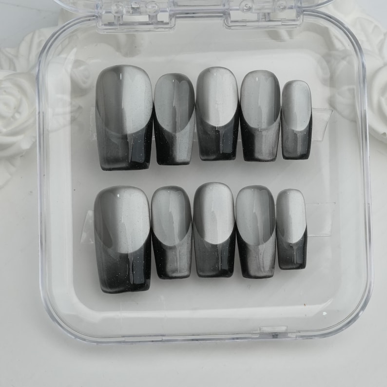 May include: A set of ten artificial nails in a clear plastic case. The nails are oval-shaped with a gradient design, transitioning from light gray to dark gray. The nails are arranged in two rows of five.