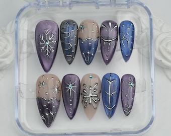 May include: A set of ten artificial nails in a clear plastic case. The nails are almond-shaped and feature a variety of designs, including snowflakes, swirls, and geometric patterns. Colors include purple, blue, and beige, with silver accents and rhinestones.