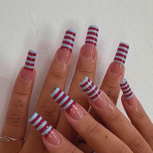 Könnte beinhalten: Eine Hand mit langen, quadratischen Nägeln. Jeder Nagel ist mit abwechselnden horizontalen Streifen in Rot und Hellblau bemalt. Die Nagelbasis hat ein natürliches, klares Finish.