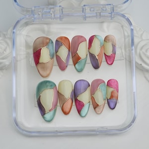 Op de afbeelding: Een set van tien kunstnagels in een doorzichtige plastic doos. Elke nagel heeft een glas-in-loodontwerp met gouden contouren en een verscheidenheid aan kleuren, waaronder roze, bruin, groen en paars. De nagels zijn ovaal van vorm.