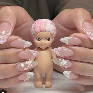 Puede incluir: Una pequeña figura de color nude con un tocado floral rosa y blanco se sostiene entre dos manos con uñas largas en forma de almendra. Las uñas están pintadas con una base rosa claro, diseños florales blancos y detalles de perlas.
